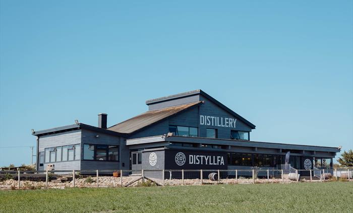 A picture of In the Welsh Wind Distillery - grey bilding with white 'distillery' 'dystyllfa' wording in front of a field of green crop